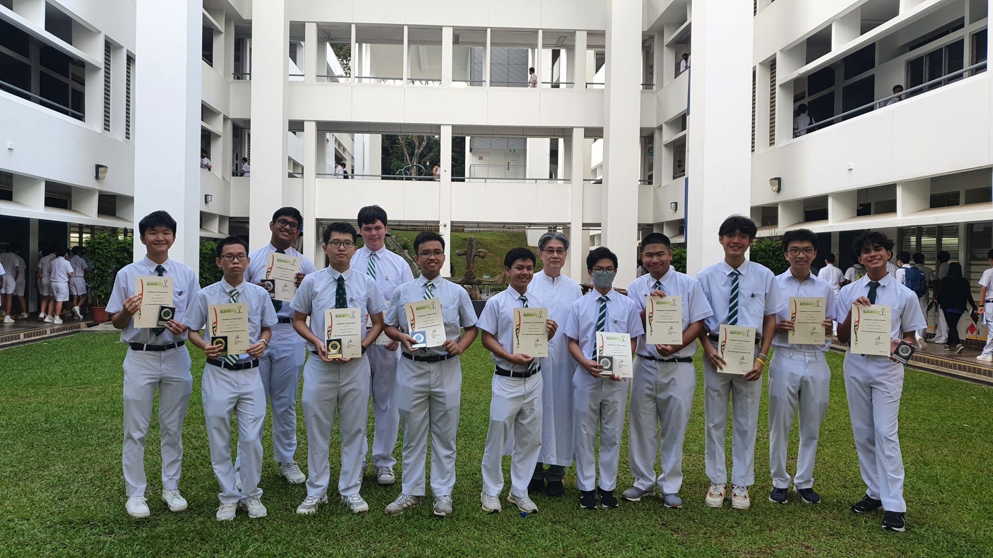 A group of male students in white uniforms holding certificates and trophies, standing on grass in a school courtyard.